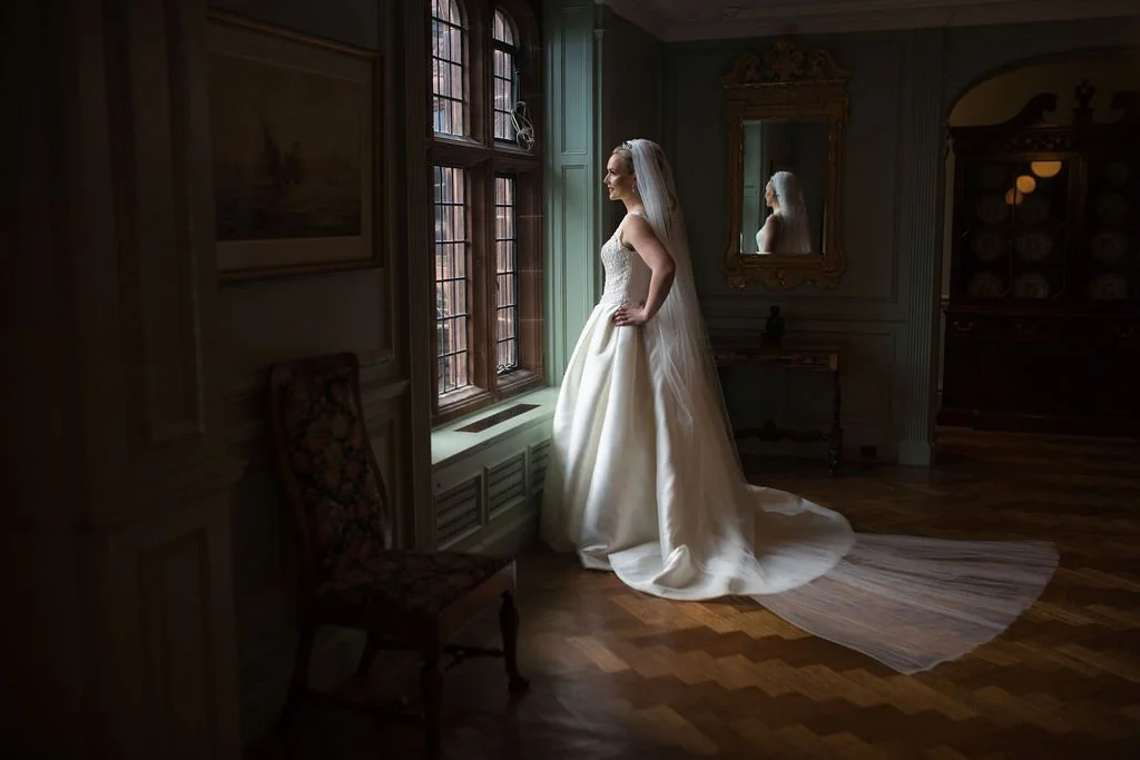 A bride in a wedding dress standing by a window in a historic room, looking out, with her reflection visible in a mirror behind her.
