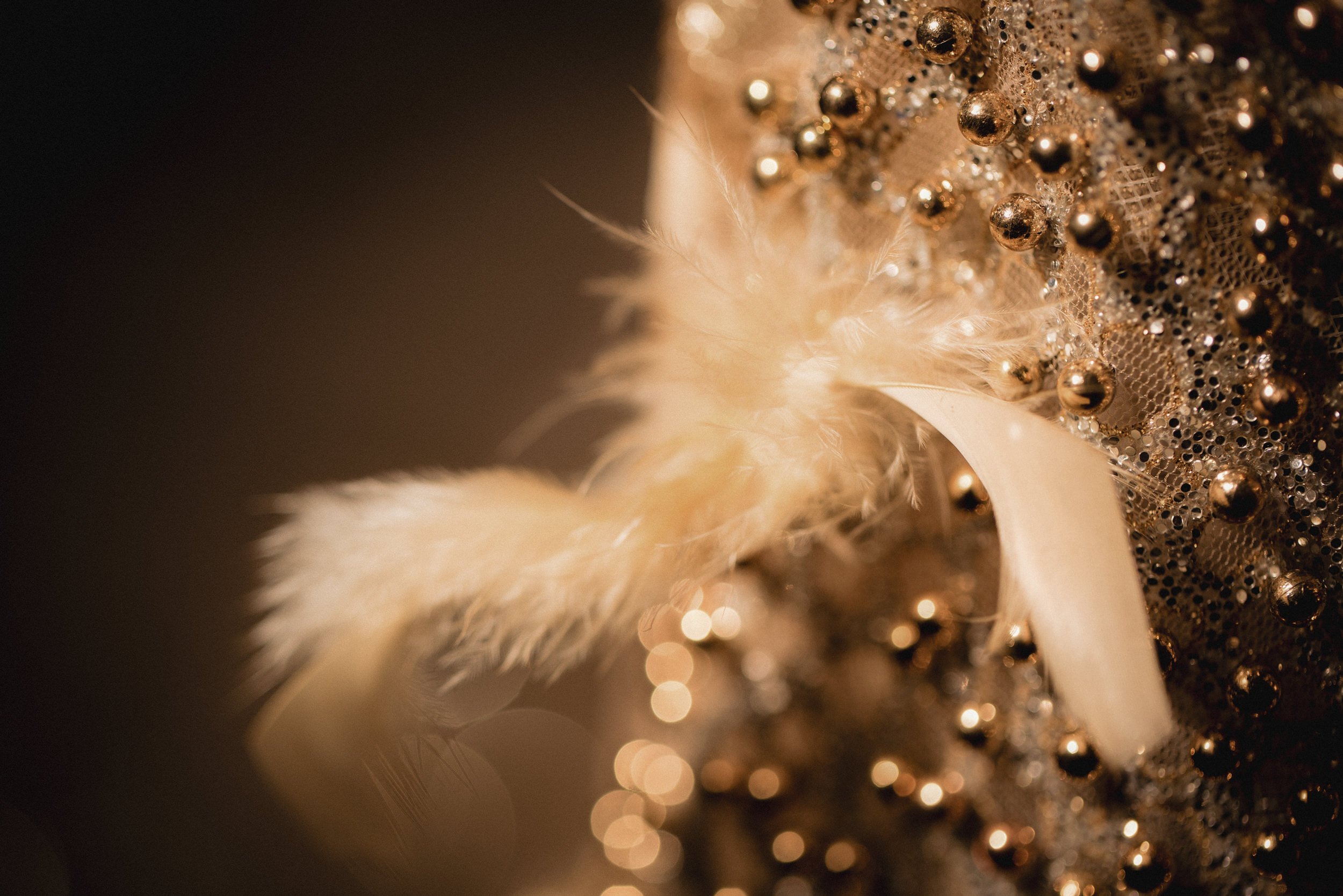 Close-up of a decorative Christmas or holiday ornament with gold beads, lace, and feathers, with warm lighting creating bokeh effects in the background.