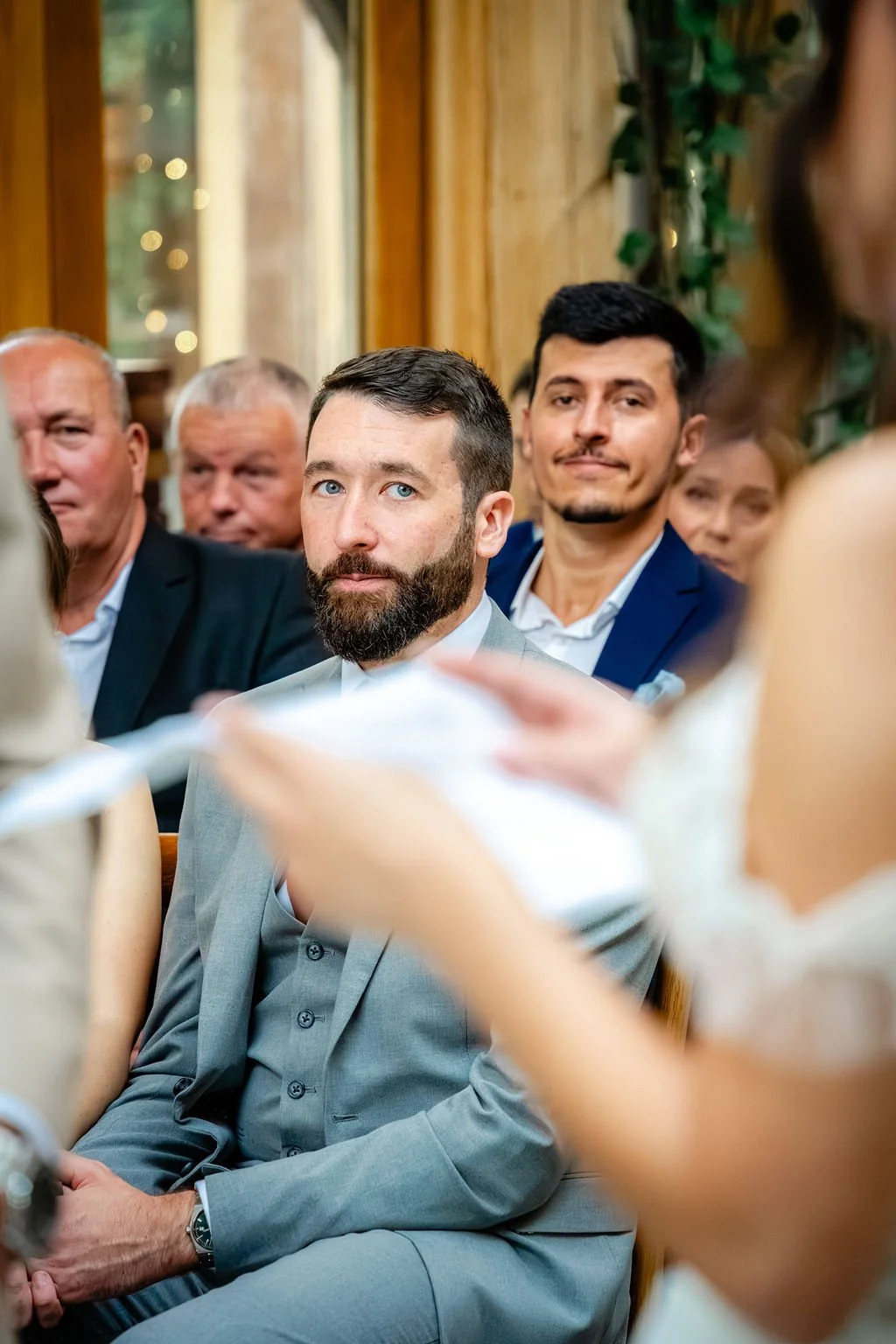 A group of people seated indoors at a formal event, with focus on a bearded man in a gray suit and a man with dark hair and a blue suit behind him. Other attendees are visible in the background.