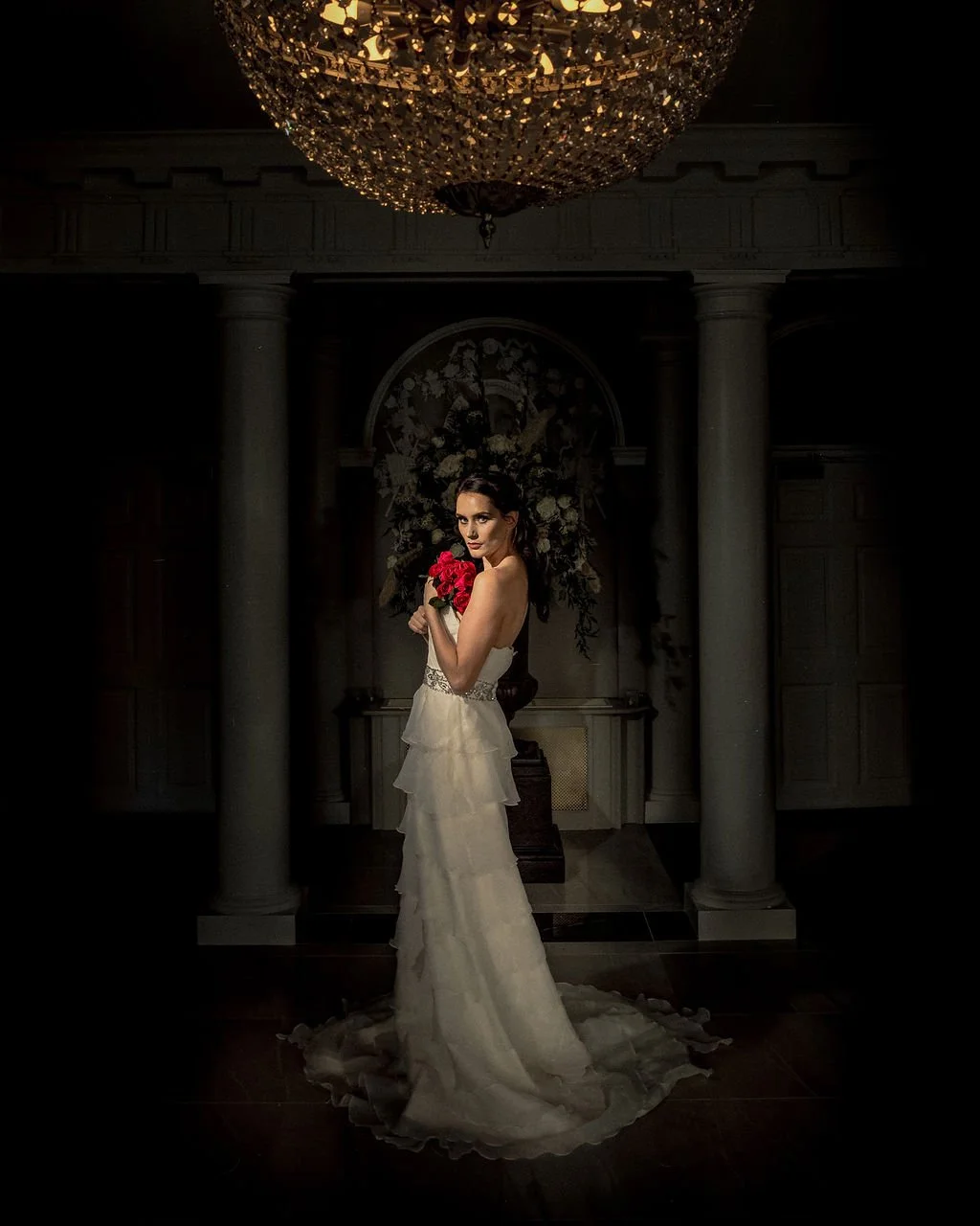 A bride in a strapless white wedding gown holding a bouquet of red roses, standing in a dimly lit room beneath a large elaborate chandelier.