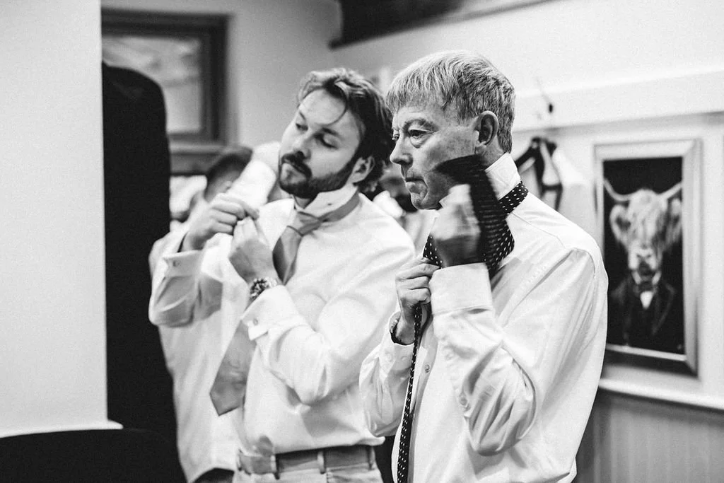 Two men dressed in white shirts are adjusting their neckties and bowties in front of a mirror, with one appearing to be younger and the other older. There is a framed picture of a bull on the wall behind them.