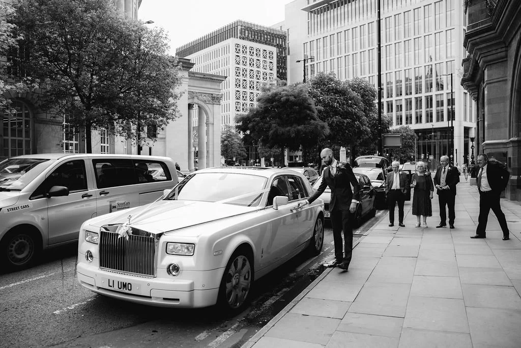 A groom in a suit is placing a key into a luxury white limousine with a ribbon on the front, on a city street, surrounded by other vehicles and pedestrians.