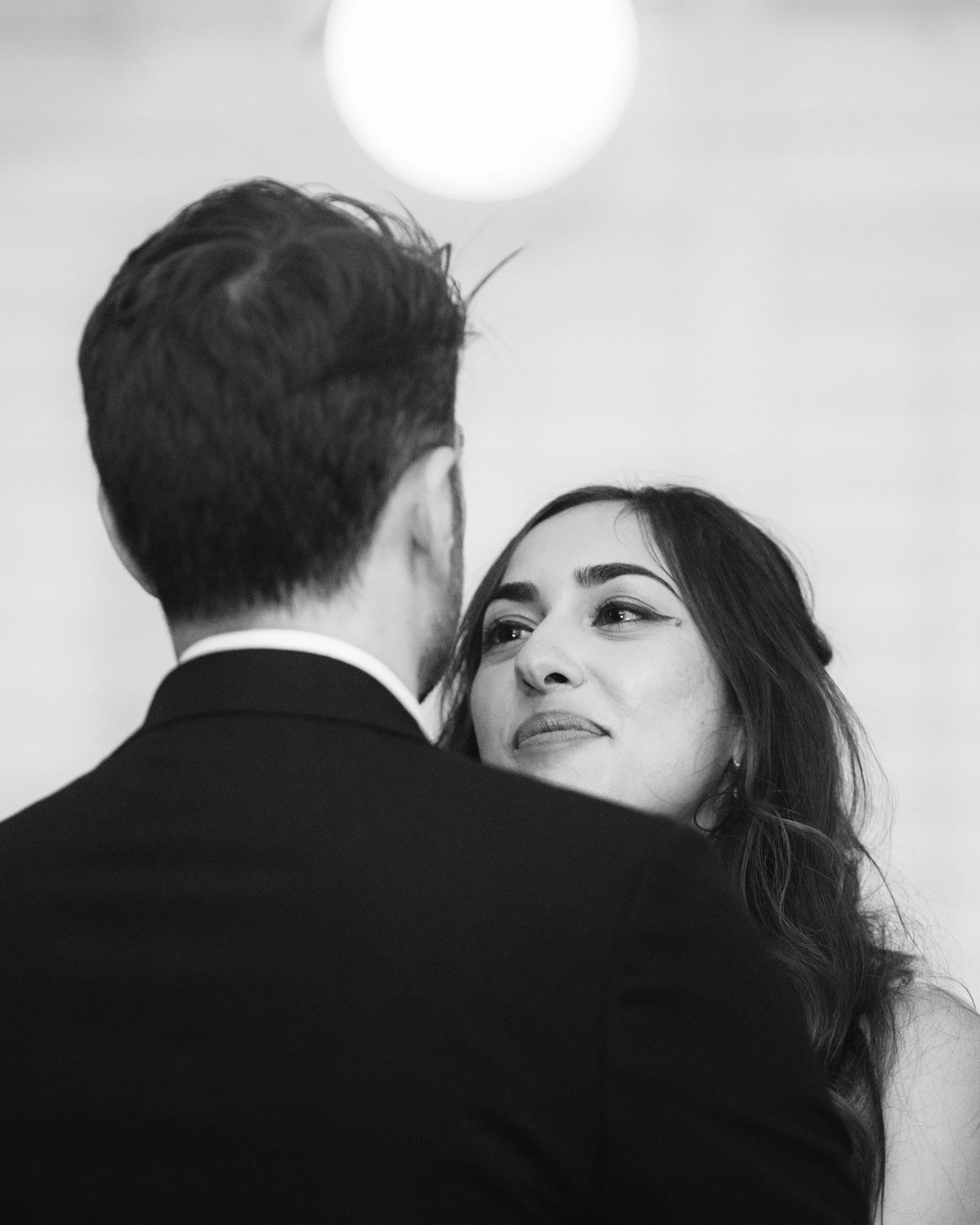 A black-and-white photo of a man and woman, possibly dancing closely, with the man's back to the camera and the woman's face visible, looking into his eyes.