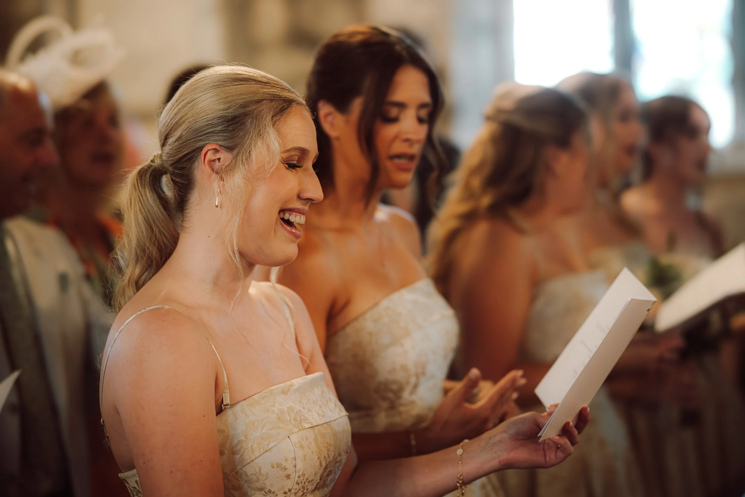 Women in formal attire attending a ceremony, reading from papers, smiling and singing.