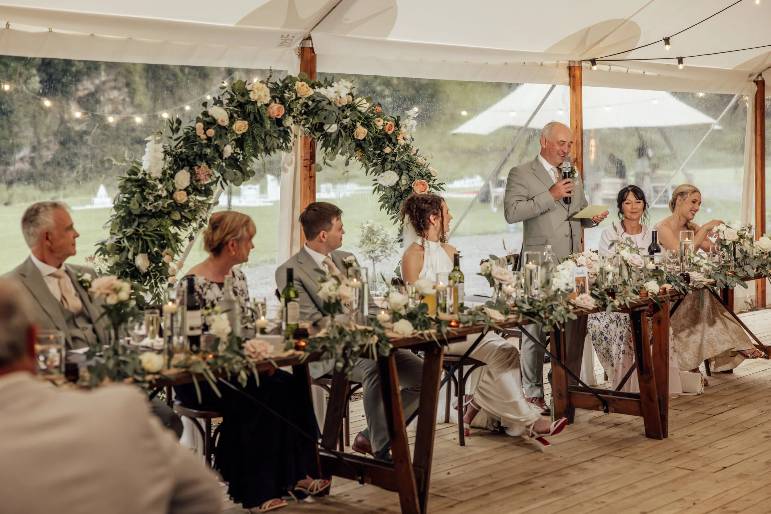 A man giving a speech at a wedding reception, standing at a decorated head table with guests seated in front, under a clear tent with string lights and a floral arch, on a rainy day by a lake.
