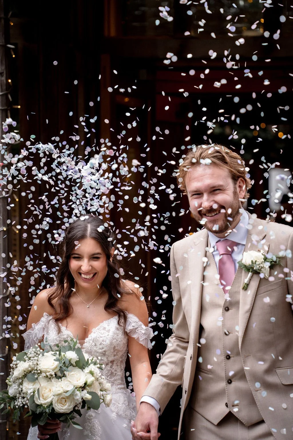 A newlywed couple is celebrating, with confetti falling around them. The bride, in a white lace wedding dress, holds a bouquet of white roses, and the groom, in a beige suit with a pink tie, smiles happily.