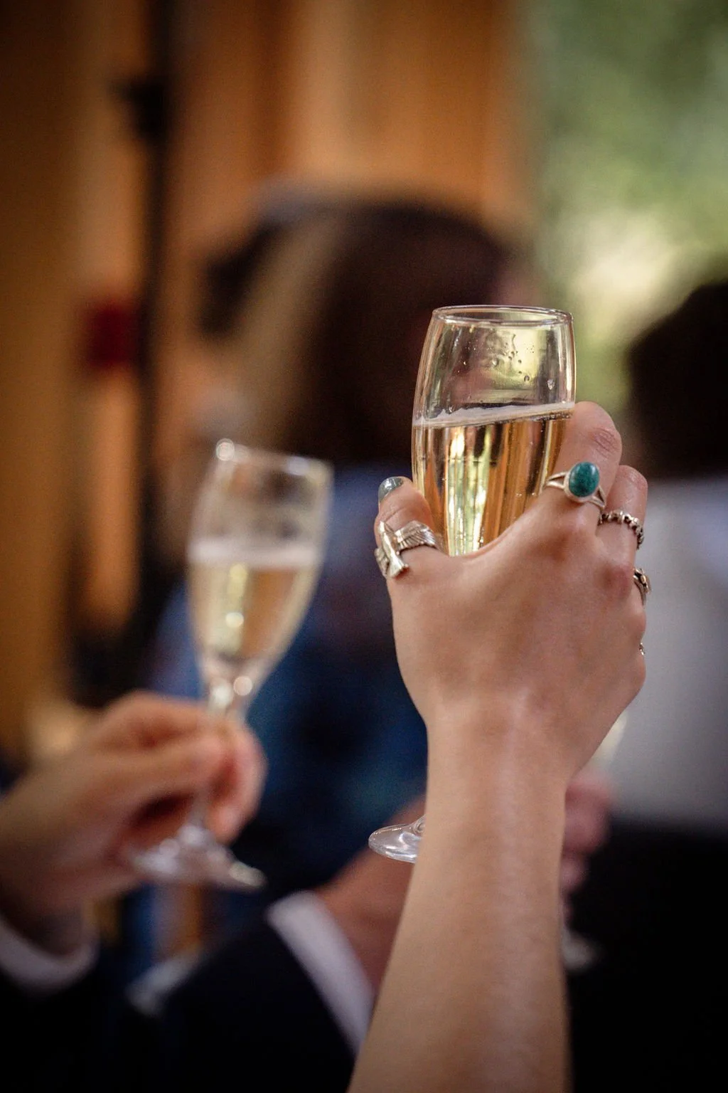 Person holding a glass of champagne with jewelry on fingers, celebrating with others in the background.