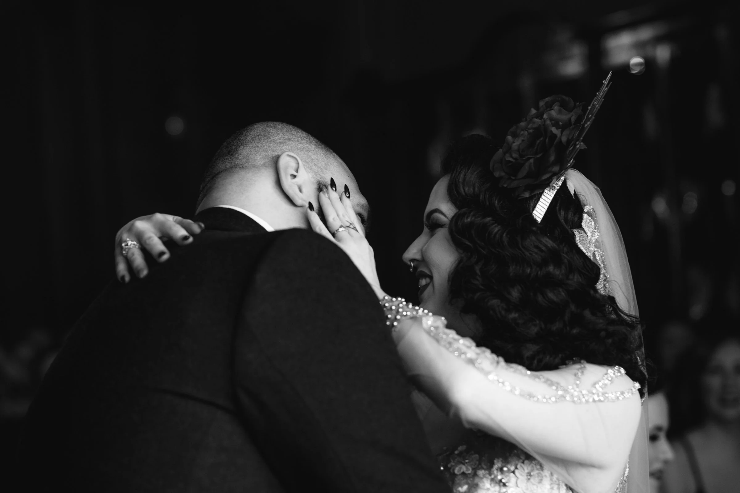 Black and white photograph of a bride and groom sharing a close, intimate moment, with the bride tenderly touching the groom's face, both smiling softly.
