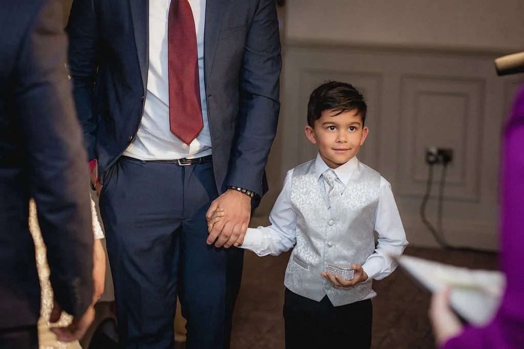 A young boy in a white vest and shirt standing beside an adult man, holding the adult's hand at an indoor event.