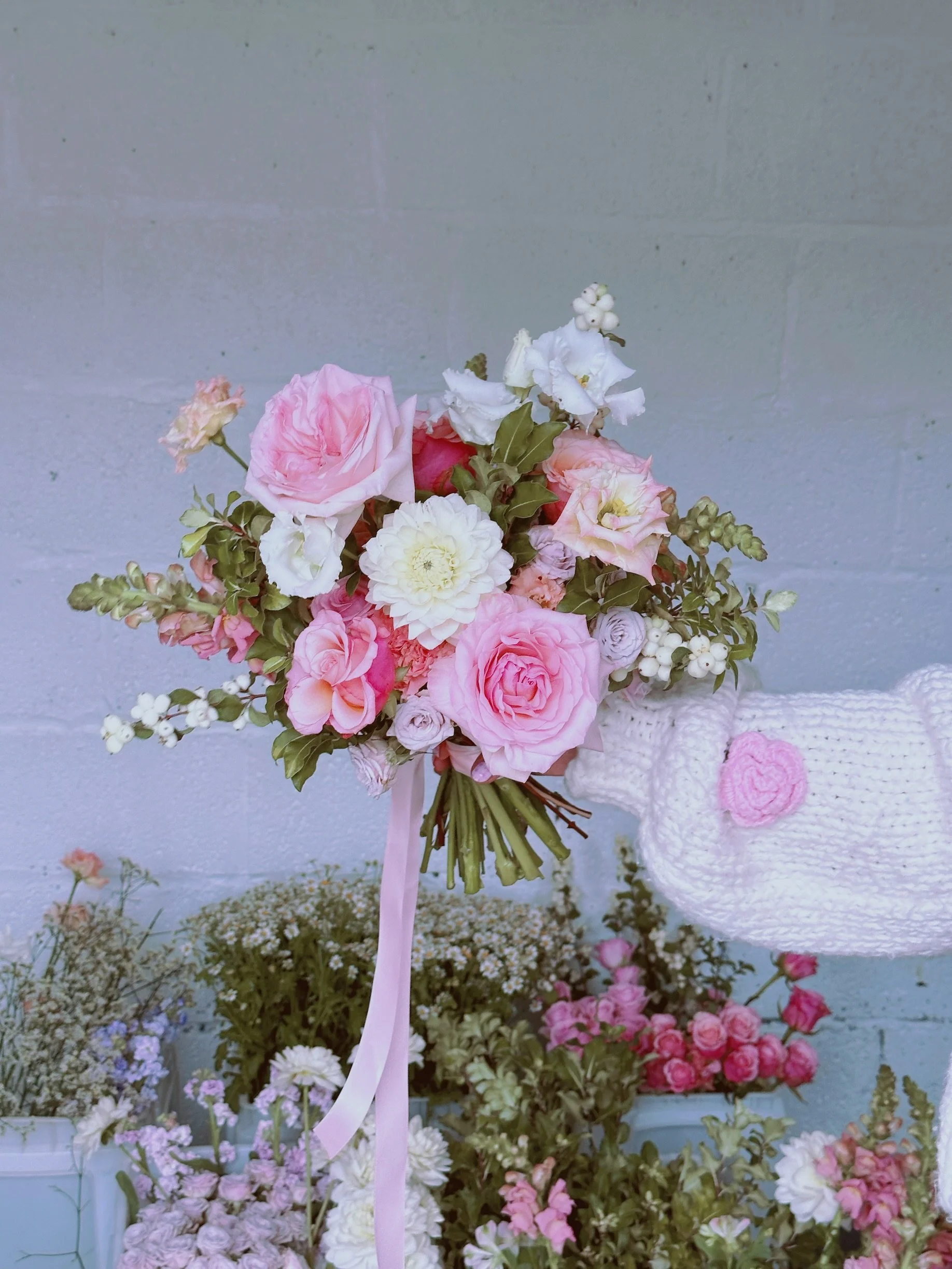 A person wearing white knitted gloves holding a bouquet of pink, white, and light purple flowers against a gray brick wall. The bouquet contains roses, and other assorted flowers, arranged with green foliage, and has a pink ribbon tied around the stems.