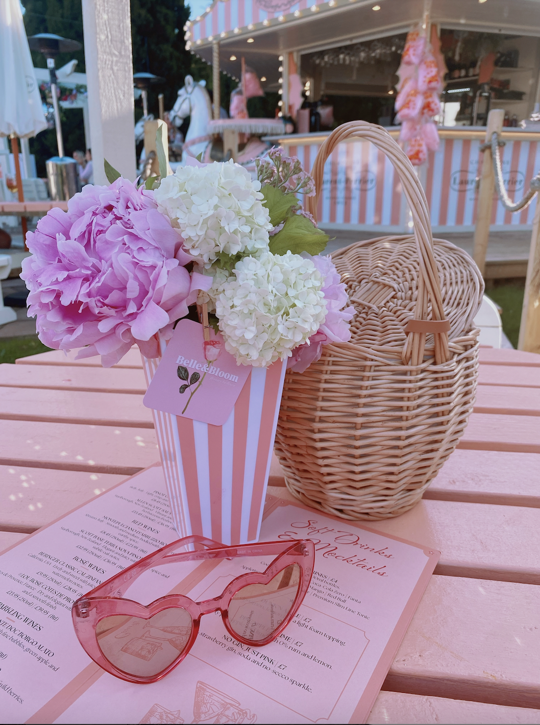 Pink and white hydrangea flowers in a pink and white striped vase on a pink outdoor table, with a pink gift card attached, pink sunglasses, a wicker basket, and a pink menu. In the background, there is a pink and white striped carousel and a bar area decorated with pink and fluffy items.