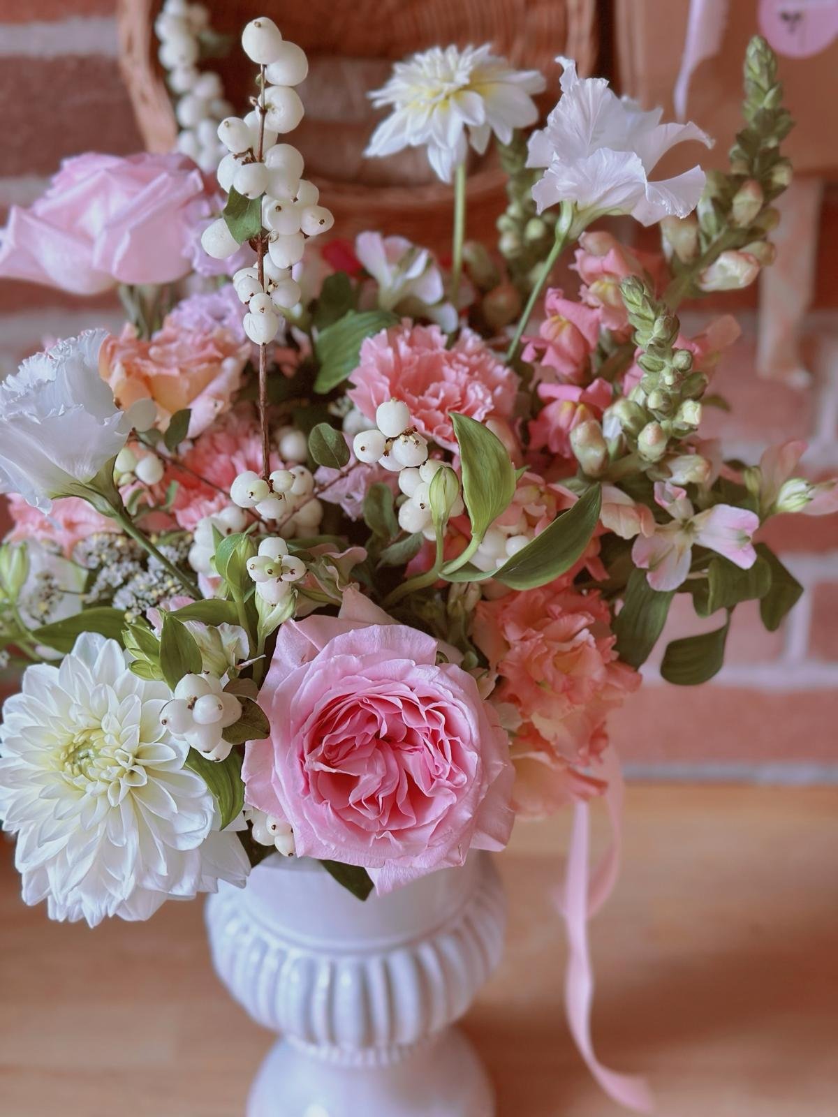 Pink and white mixed flower bouquet in a white vase, including roses, carnations, dahlias, and berries, on a wooden surface with a brick wall background.