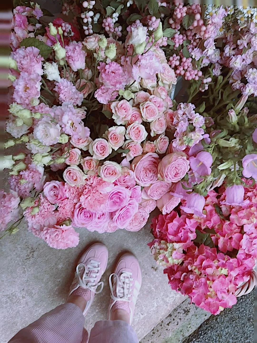 A display of various pink and white flowers, including roses and peonies, with part of a person's feet in pink sneakers and striped pants visible at the bottom.