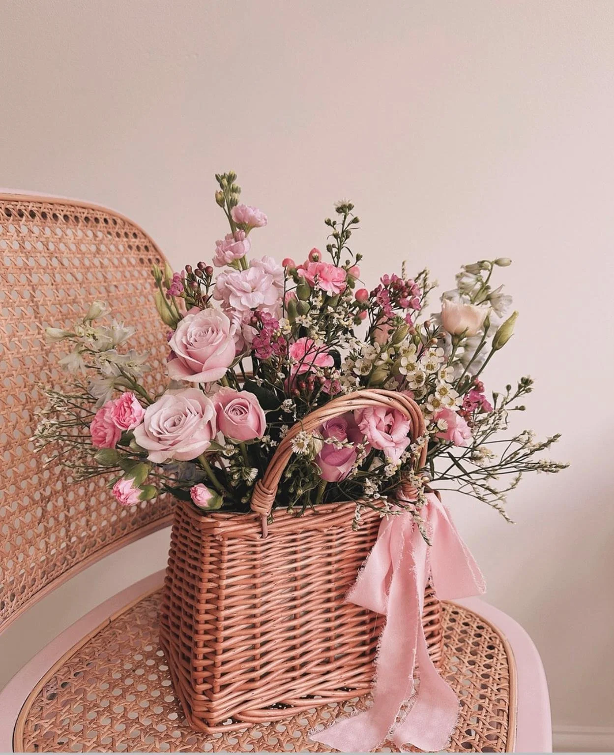 A pink wicker basket filled with pink and white flowers, including roses, arranged on a pink rattan chair.