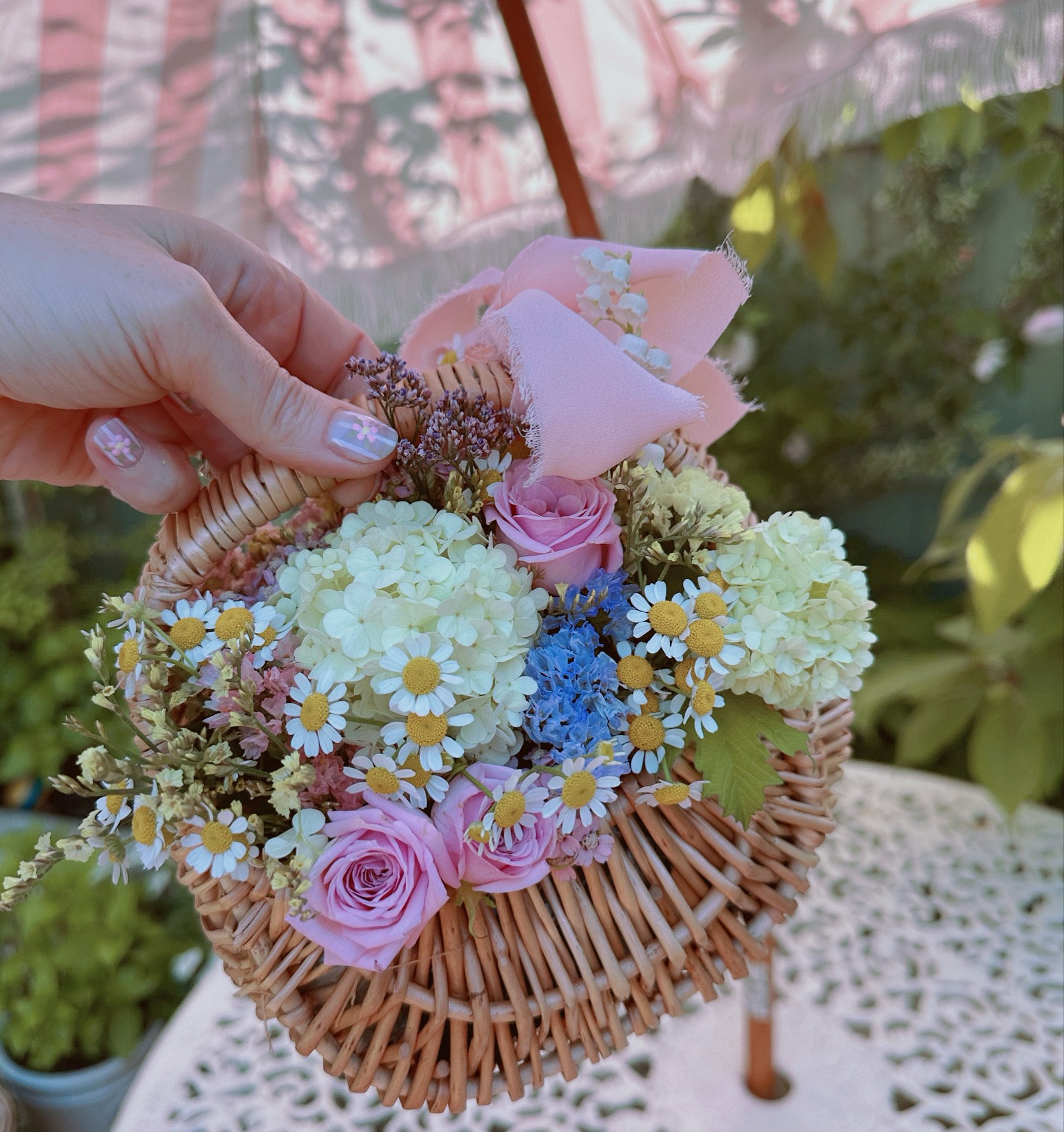 A hand holding a woven basket filled with pink roses, white hydrangeas, small daisies, blue flowers, and pink fabric ribbons, outdoors.