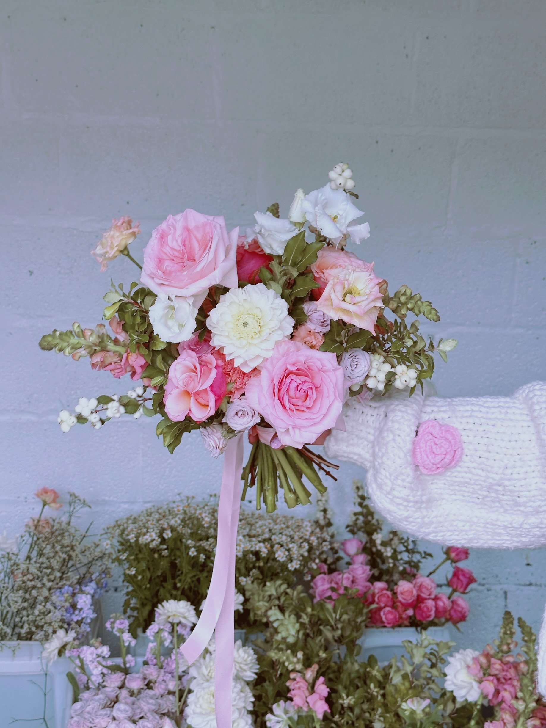 A bouquet of pink and white flowers held by a person wearing white mittens with a pink heart, against a light blue wall background.