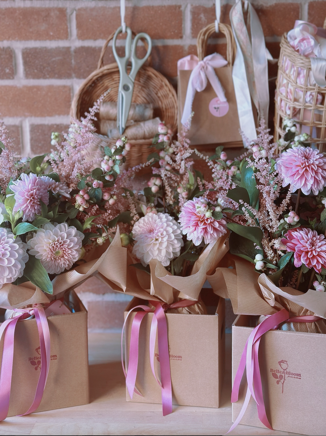 Pink floral bouquets wrapped in brown paper and tied with pink ribbons, displayed on a light wooden surface in front of a brick wall.