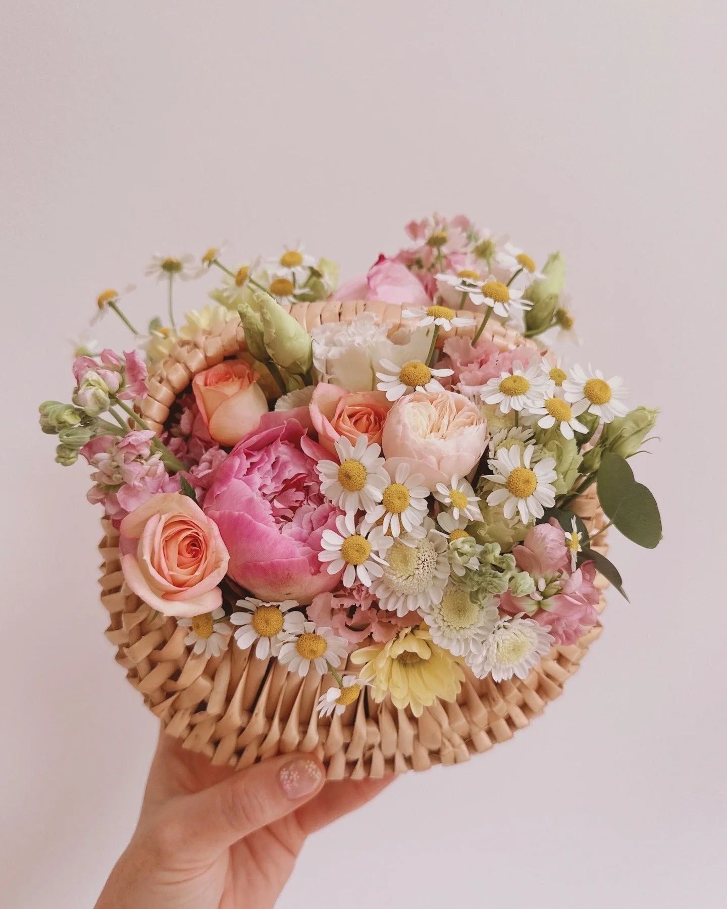 A hand holding a floral arrangement in a round woven basket, featuring pink roses, white daisies, and other pastel-colored flowers against a plain white background.