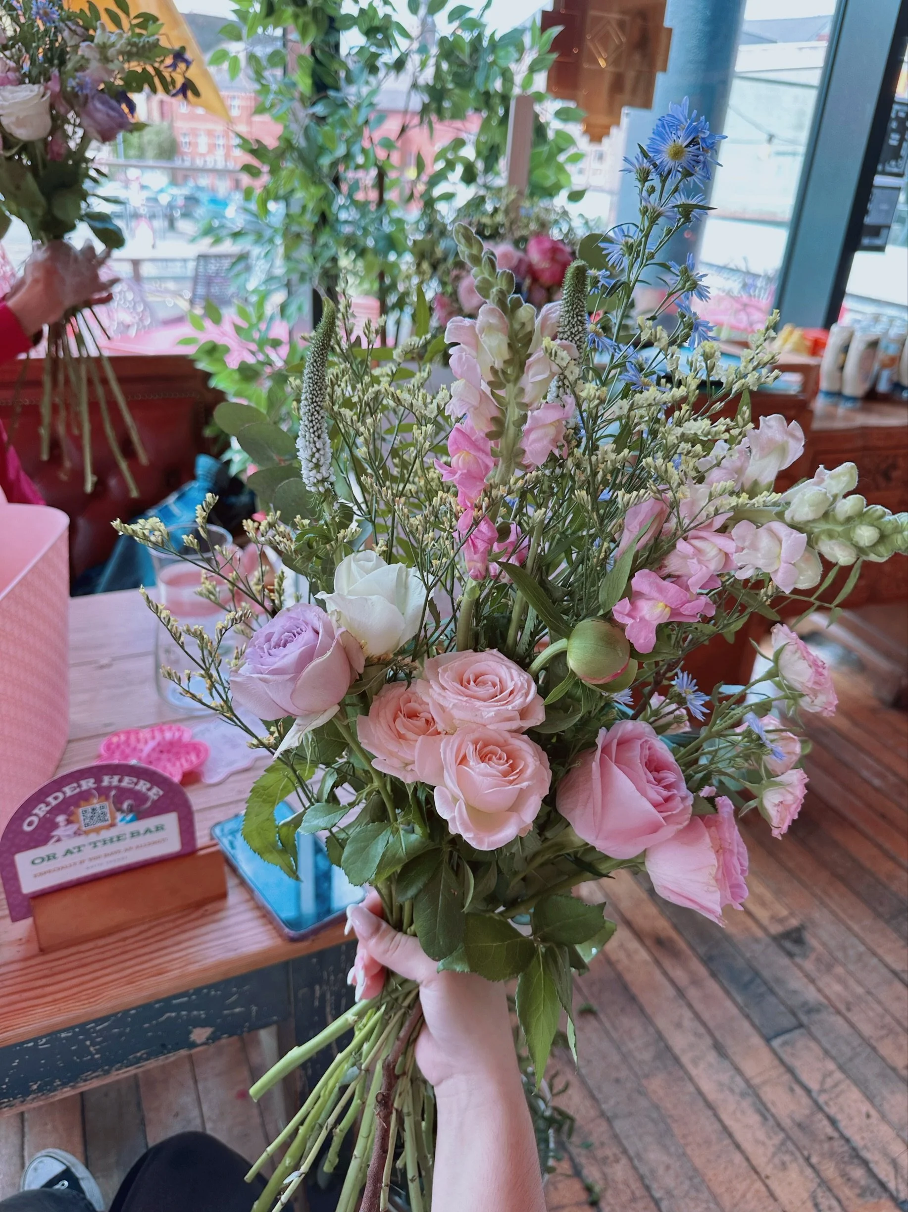 A hand holding a bouquet of pink and white roses, blue and pink flowers, and greenery inside a flower shop with wooden floors and a window view of buildings outside.