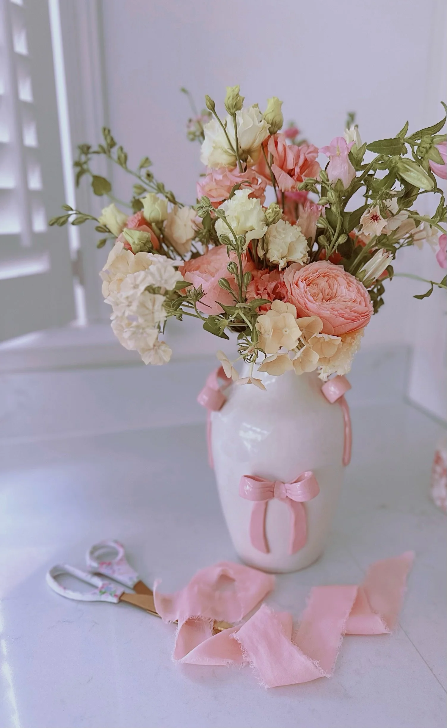 A white vase with pink ribbons holds a bouquet of pink and white flowers. Flower scissors and pink ribbon scrolls are on the table.