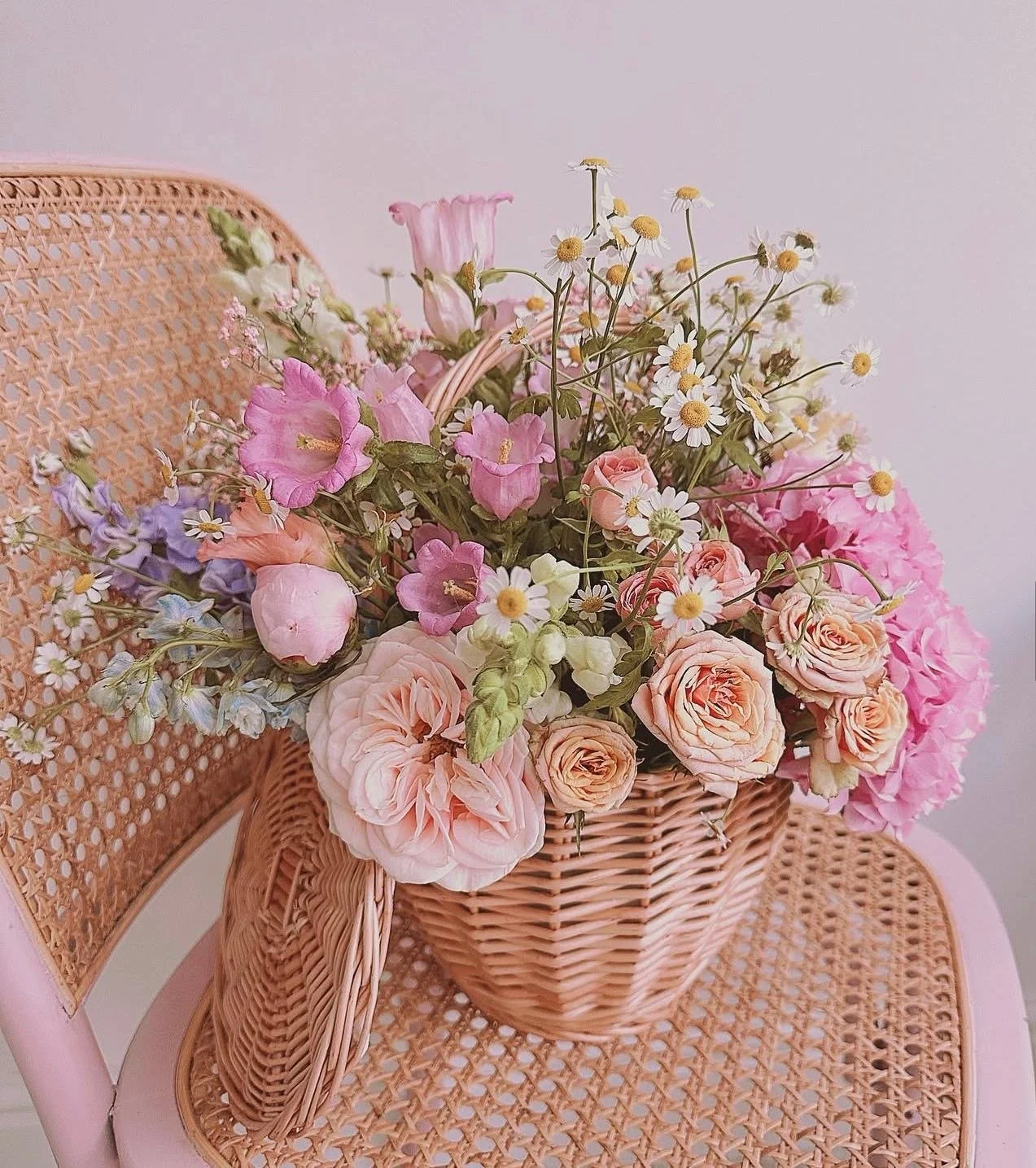 A basket filled with pink and white roses, purple and pink lisianthus, daisies, and other mixed flowers, resting on a pink woven chair.