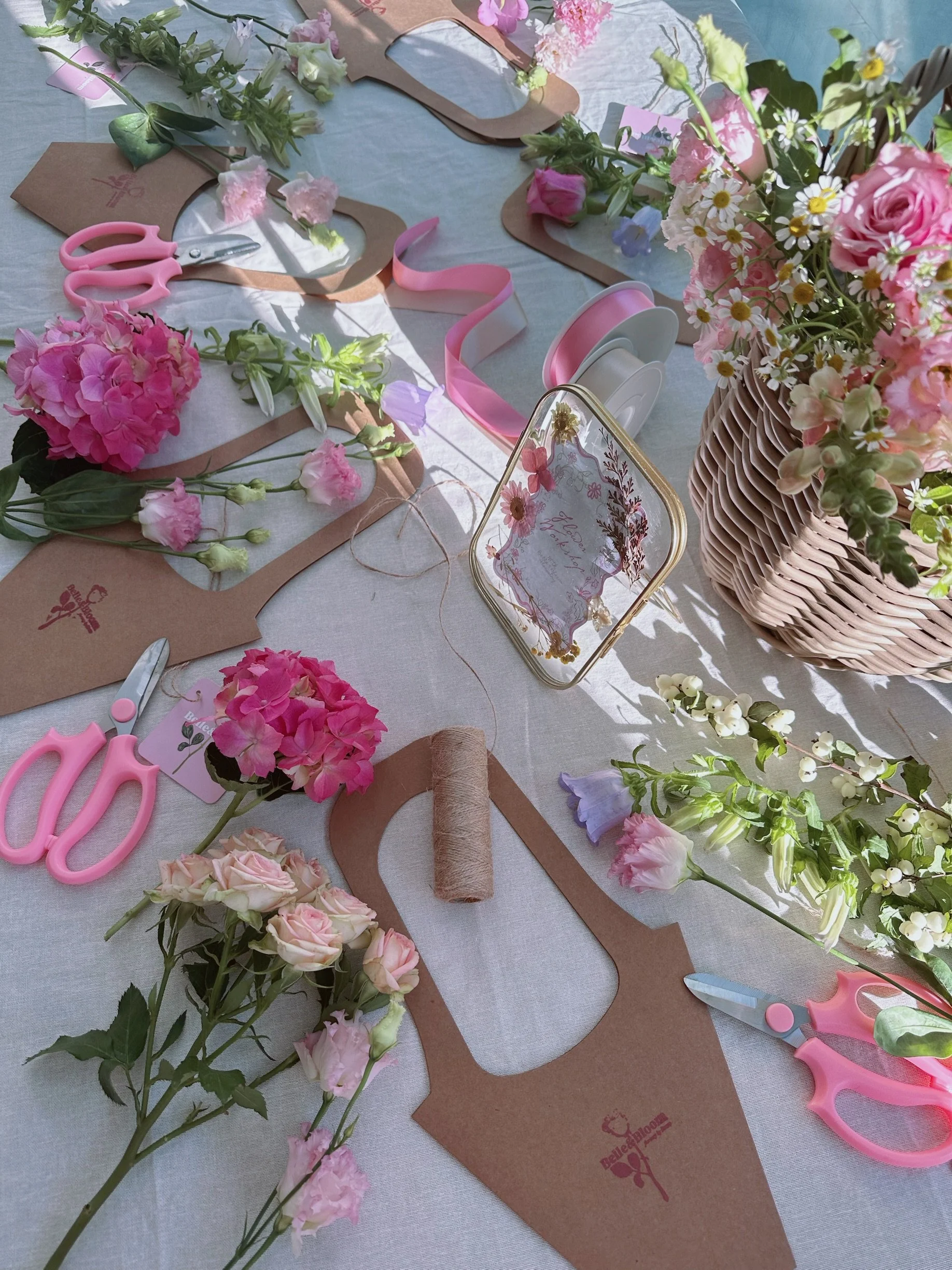 Floral arrangement and craft materials on a table, including pink flowers, scissors, ribbon, cardboard cutouts, and a wicker basket with pink and white flowers.