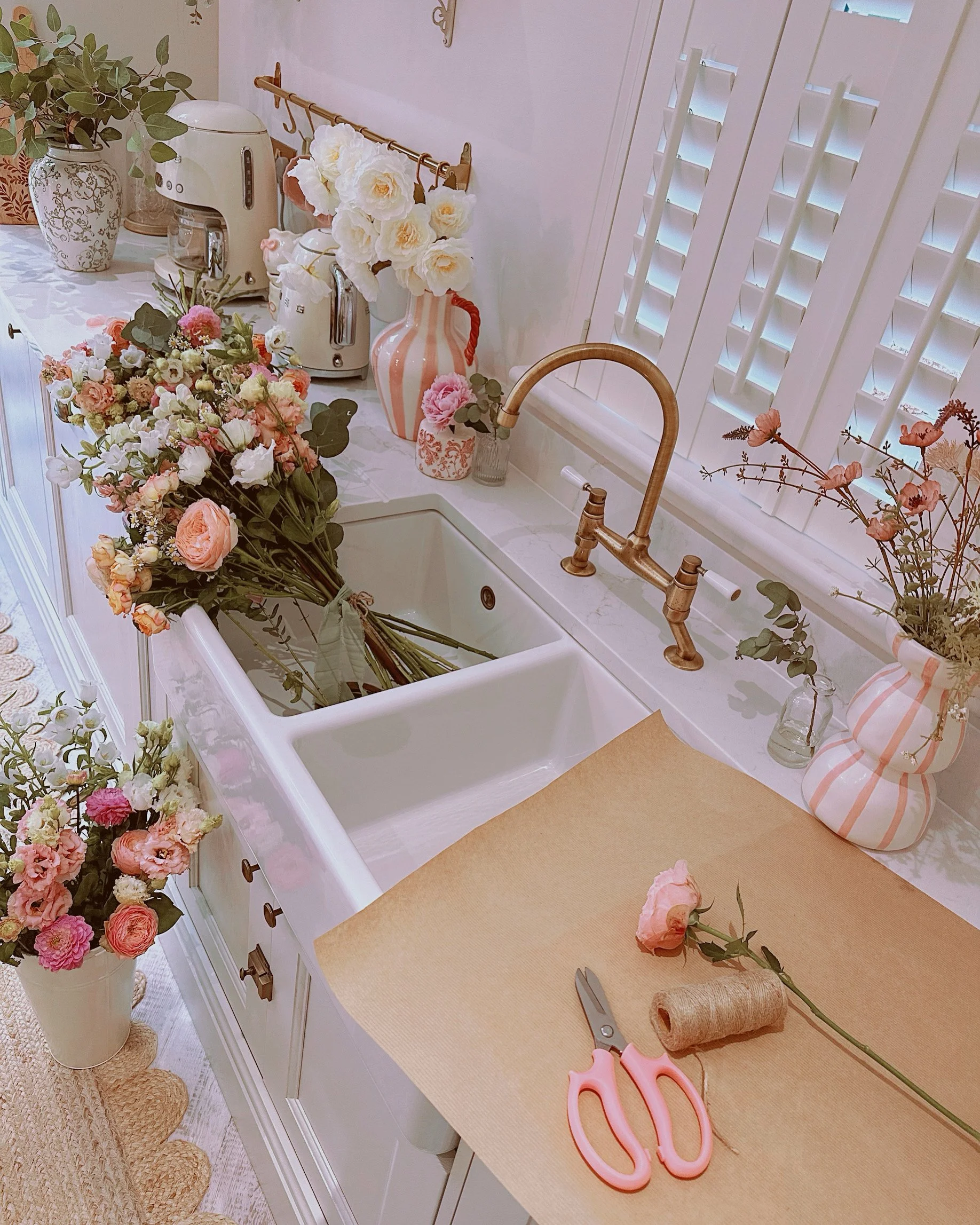 A garden-style farmhouse kitchen sink decorated with pink, white, and peach flowers in vases, with a bouquet of flowers on the counter. The window has white shutters, and there is a rose in a small jar. Pink scissors and a spool of twine are on the p