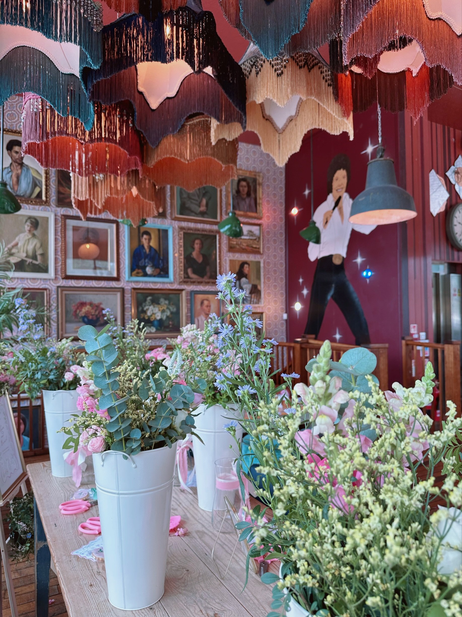 A cozy room decorated with floral arrangements in white vases, surrounded by portraits on the wall, with hanging fabric art pieces and artwork of Elvis Presley on a red wall.
