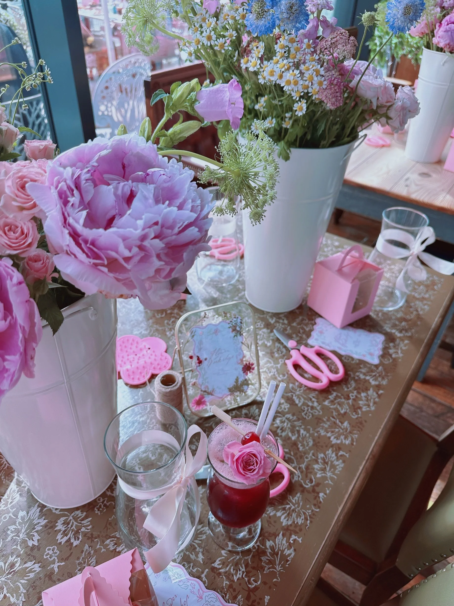 A decorated table with pink and white floral arrangements, pink scissors, a pink gift box, a glass of water with a ribbon, and a drink garnished with a pink flower and cherries, set in a cozy room with windows and natural light.