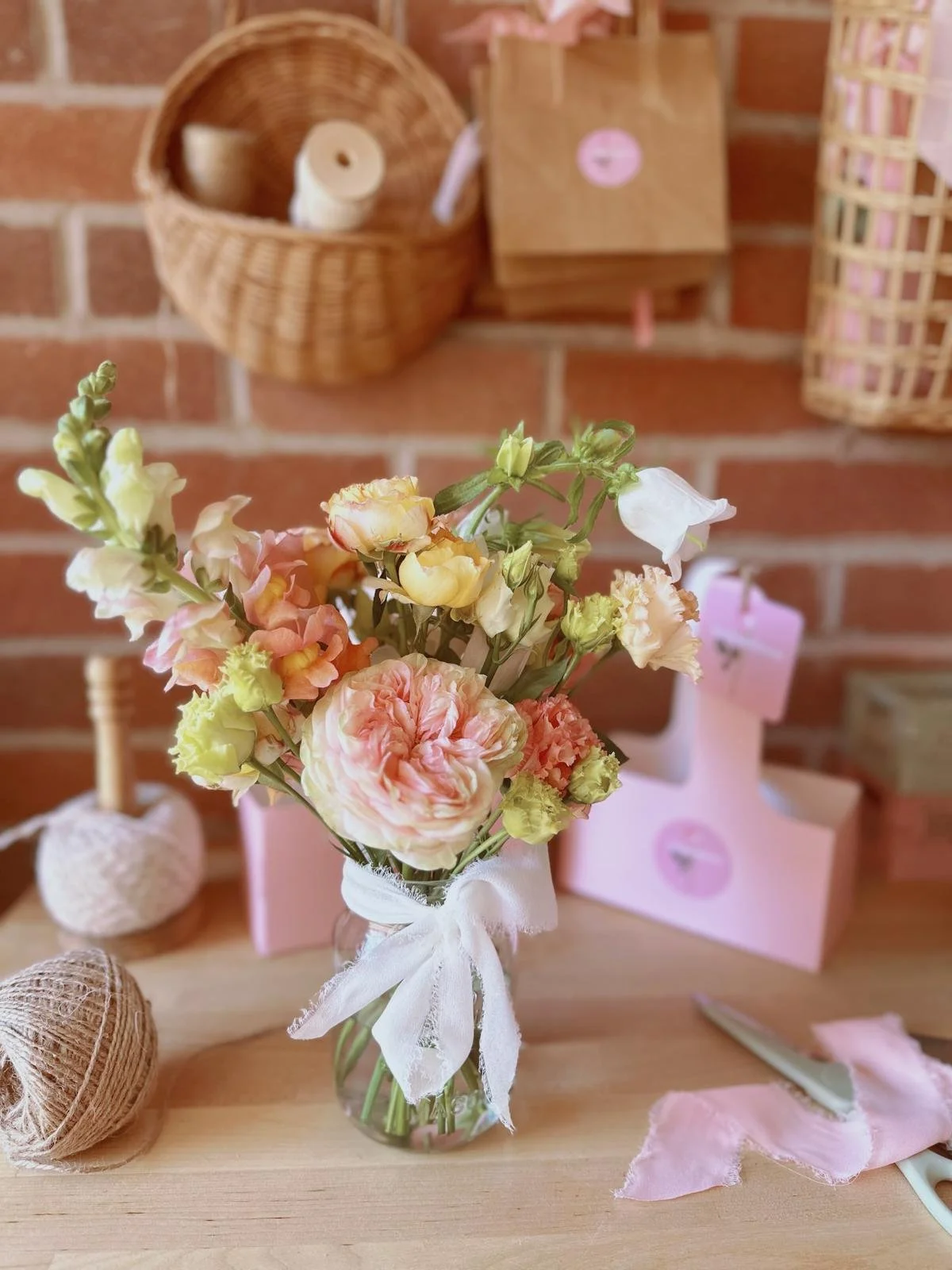 A vase with pink, peach, and white flowers tied with a white ribbon, placed on a wooden table, with craft supplies and wicker baskets in the background.