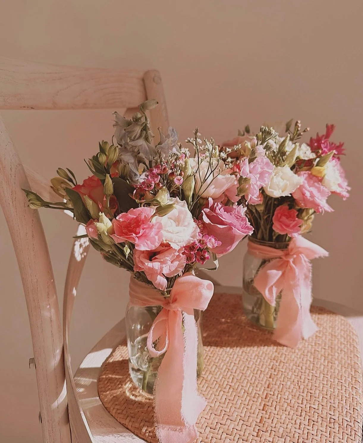 Two glass jars filled with pink and white flowers, each tied with a pink ribbon. The jars are on a woven placemat on a wooden chair.