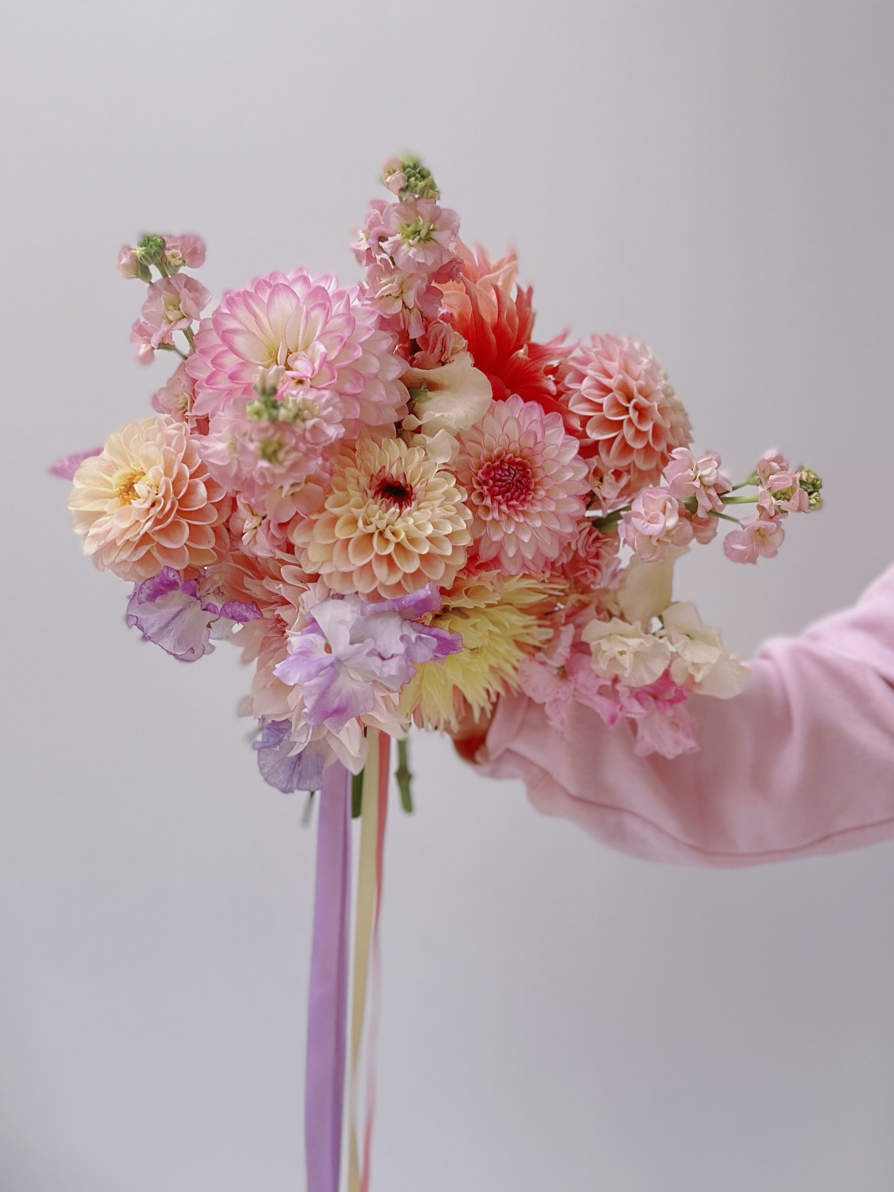 A hand in a pink glove holding a colorful bouquet of various pink, peach, cream, and purple flowers against a plain light background.