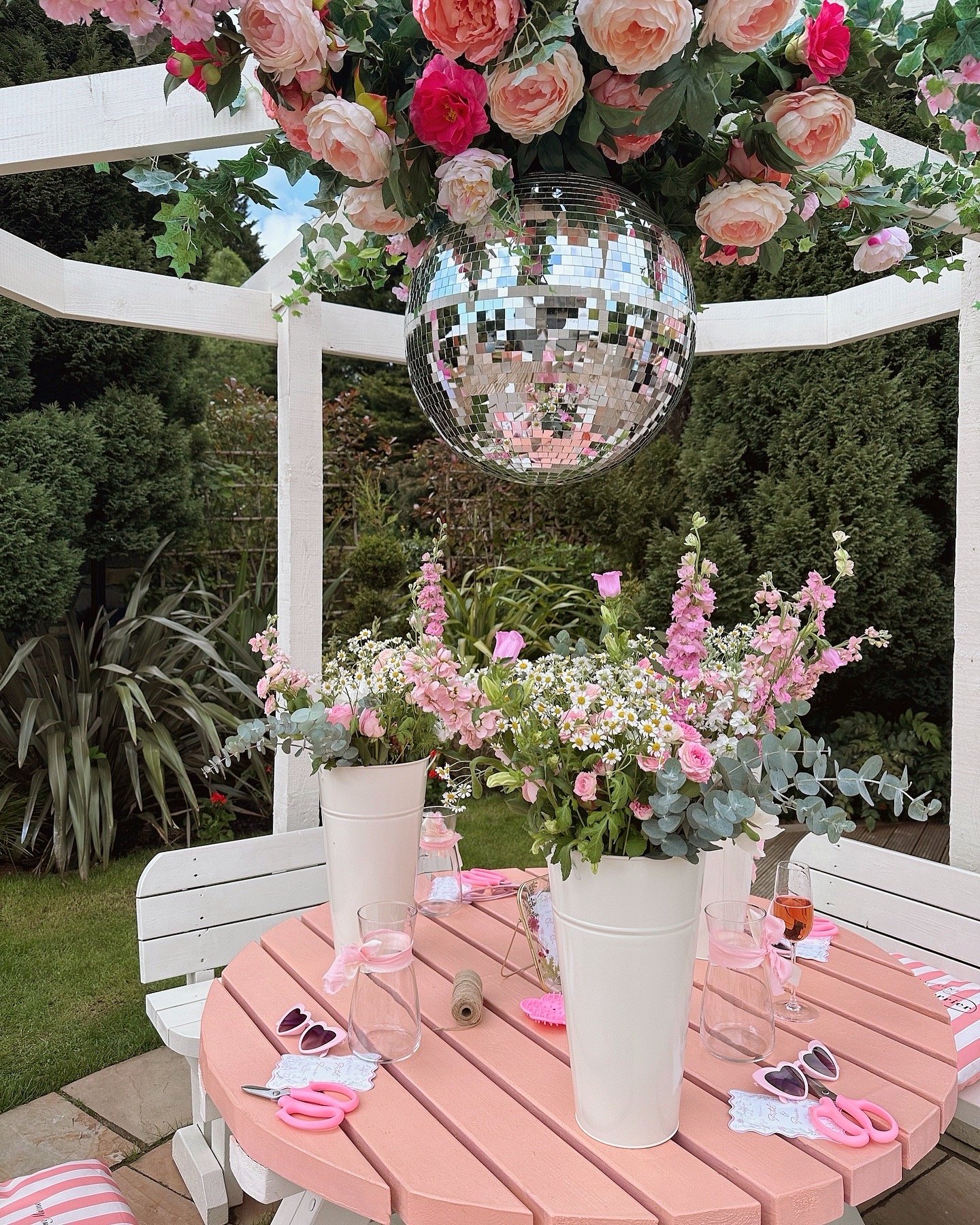 Outdoor garden table decorated with pink flowers and party accessories, under a large disco ball and floral canopy.
