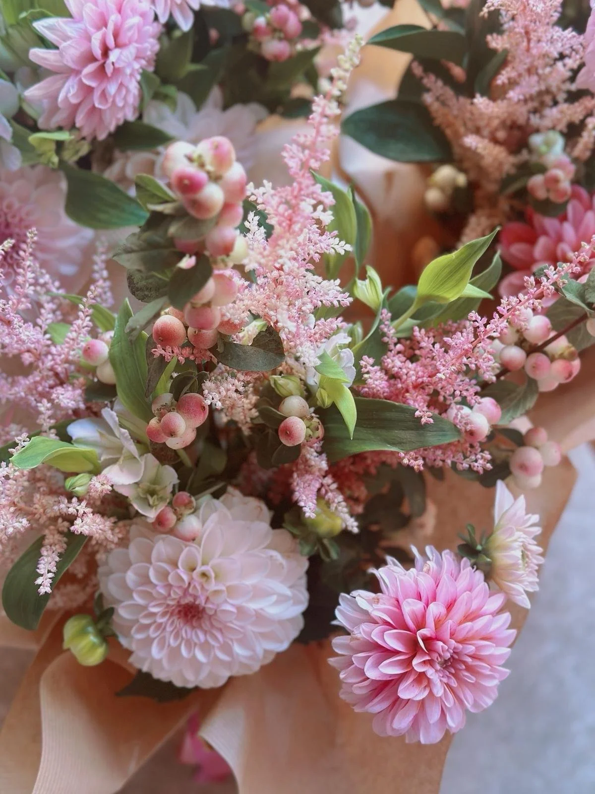 Close-up view of a bouquet of pink and white flowers, including dahlias, astilbe, and hypericum berries, wrapped in brown paper.