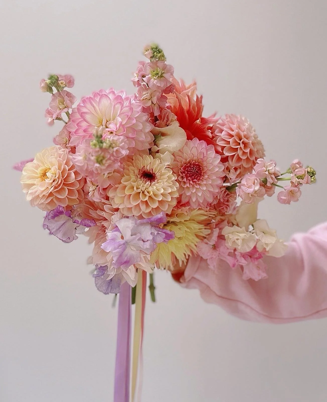 Hand holding a bouquet of pink, peach, cream, and purple flowers against a plain white background.