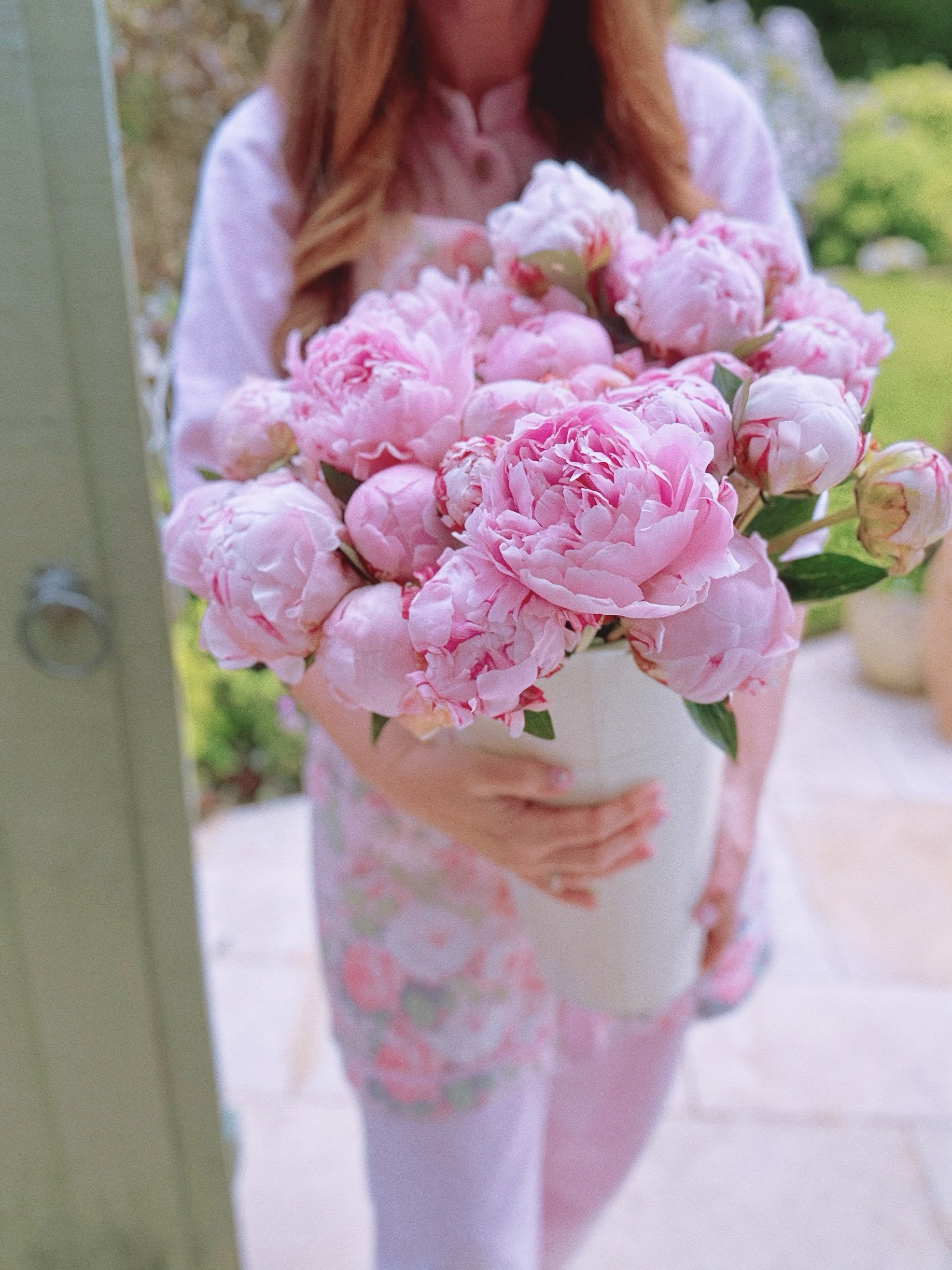 Person holding a white vase with pink peony flowers, outdoors in a garden setting.