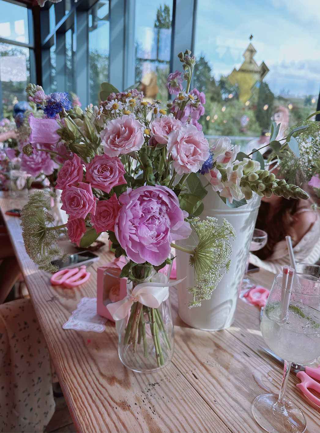 A floral arrangement with pink and purple flowers in a small glass vase on a wooden table. There are scissors, a pink gift box, a doily, and a glass of soda with a straw nearby.