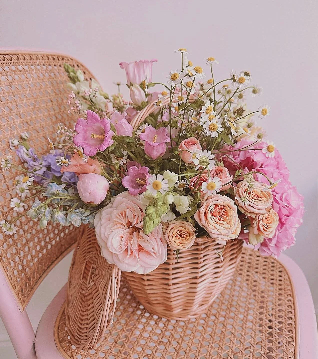 A wicker basket filled with pink, peach, purple, and white flowers placed on a pink chair with a woven backrest against a plain light background.