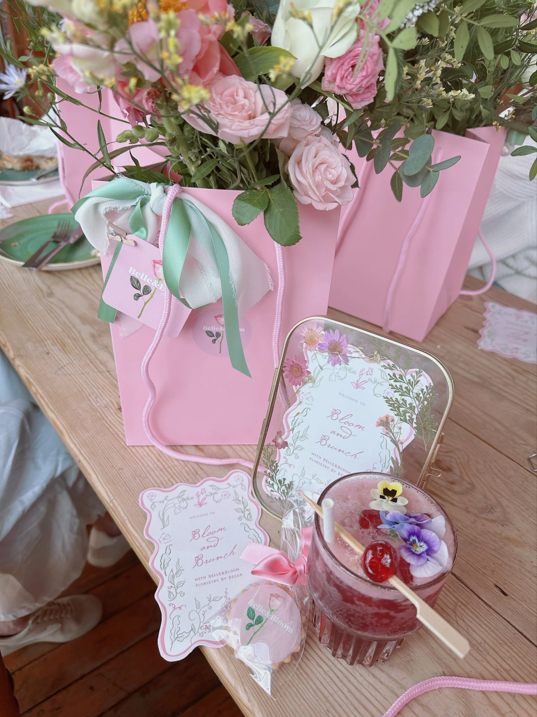 Pink gift bag with a floral arrangement and a card for a spring event, a pink drink garnished with edible flowers, cherries, and a cherry-flavored marshmallow, on a wooden table.