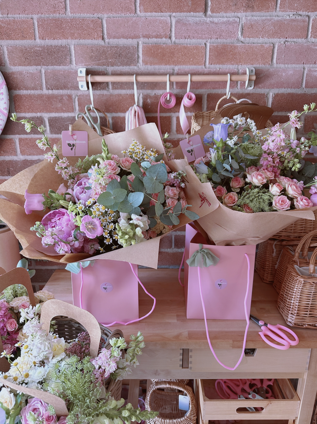 Two pink gift bags filled with assorted pink, white, and purple flowers, set on a wooden table against a brick wall. There are other floral arrangements and baskets nearby.