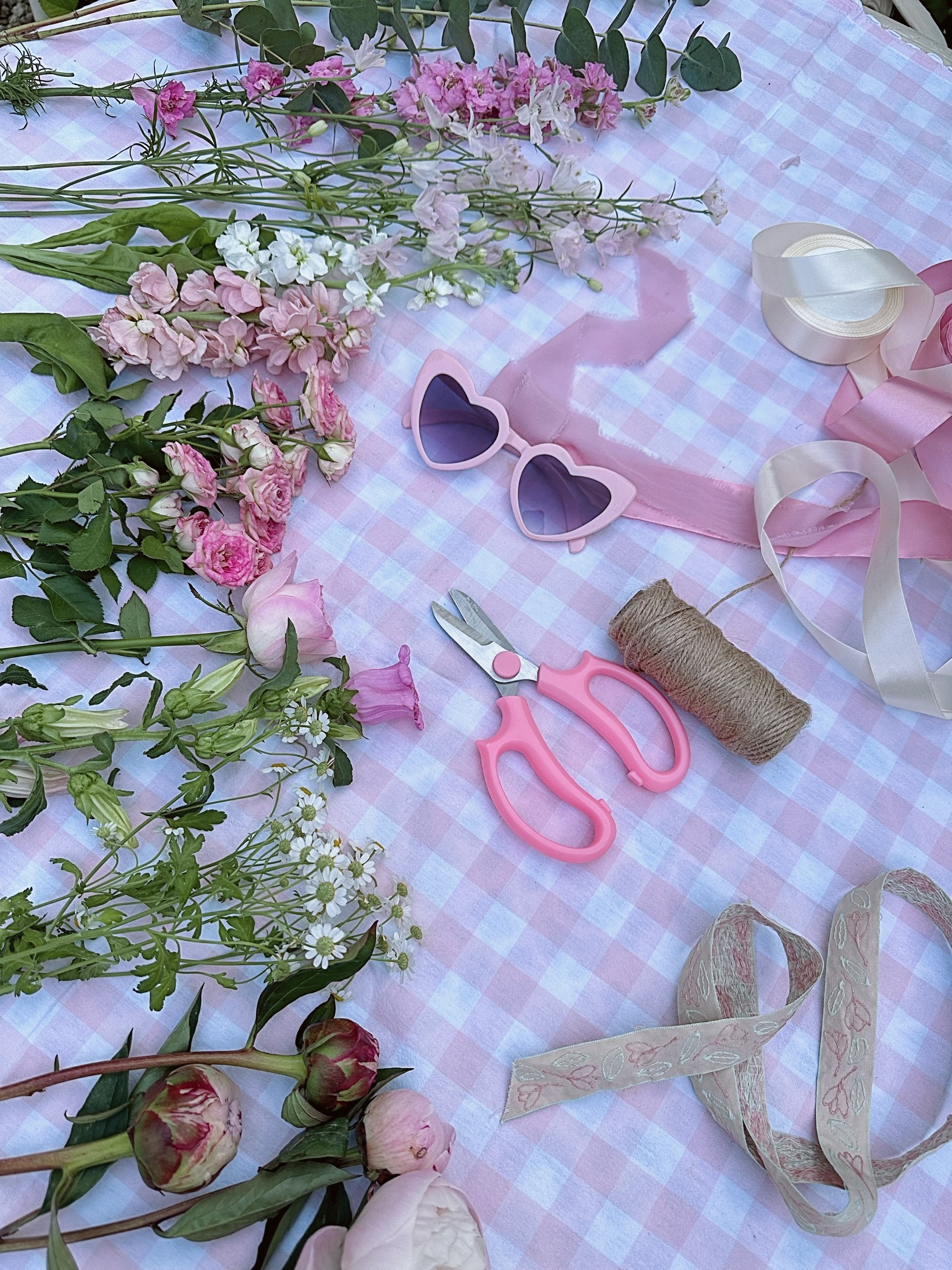 A pink and white checkered tablecloth decorated with pink and white flowers, pink heart-shaped sunglasses, pink scissors, a spool of twine, ribbons, and floral-patterned fabric.