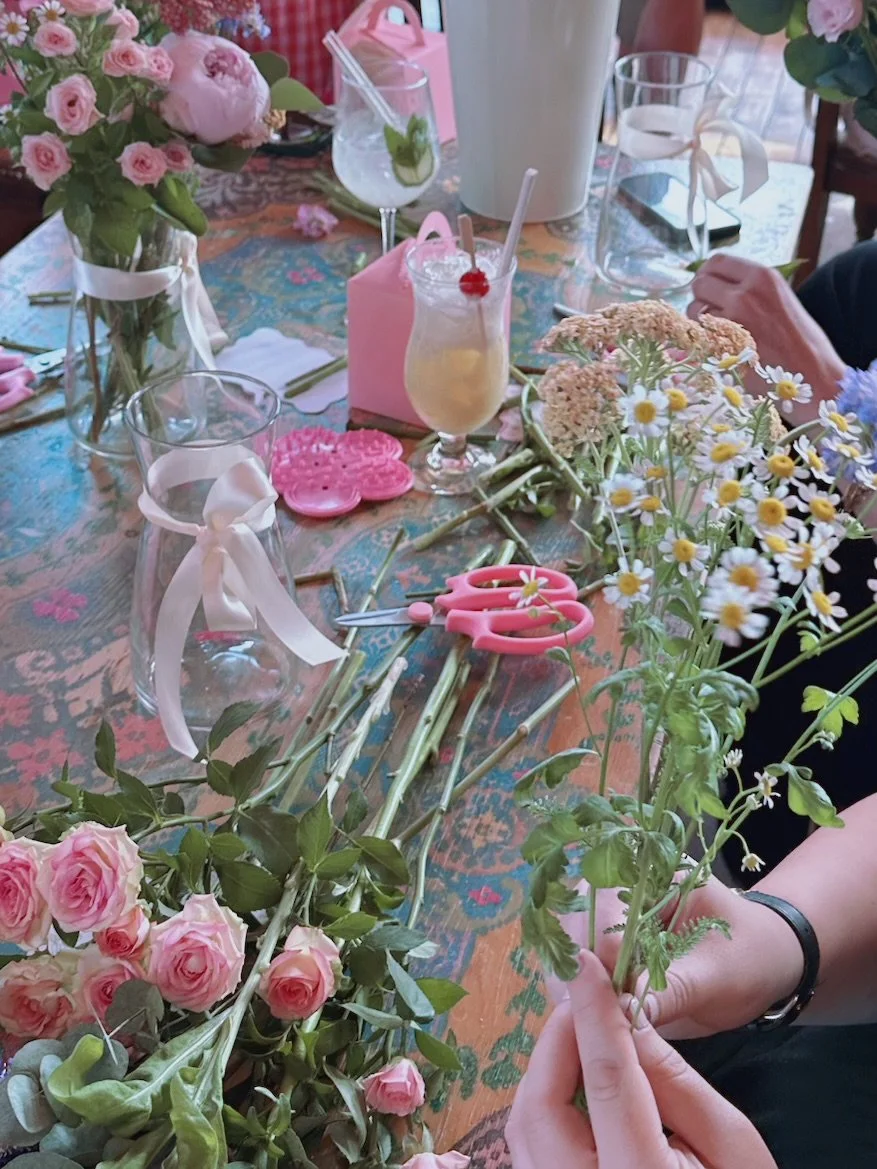 A person holding a bunch of daisies on a decorated table with flowers, scissors, drinks, and floral arrangements.