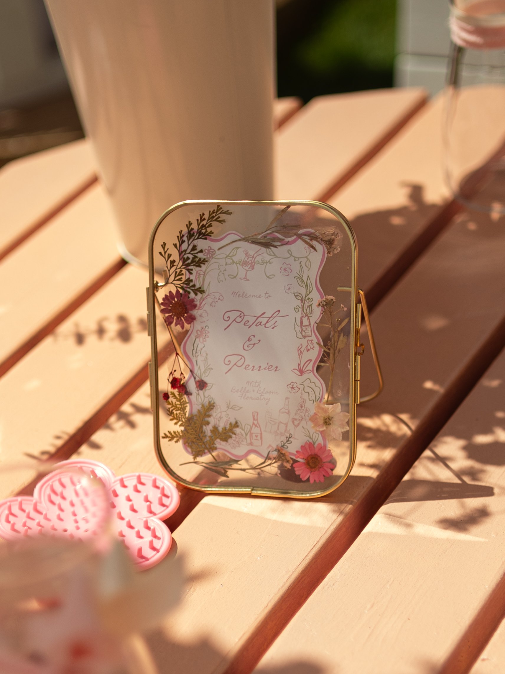 A decorative glass photo frame with floral designs and text, placed on a wooden table outdoors. The frame contains a card for an event called 'Petals & Penner' at a beauty or wellness location.