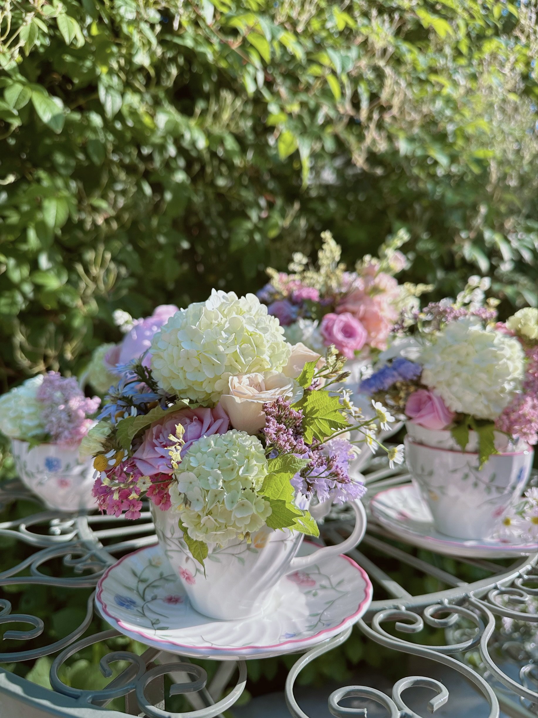 Multiple floral arrangements in patterned teacups on a decorative metal table outdoors with greenery background.