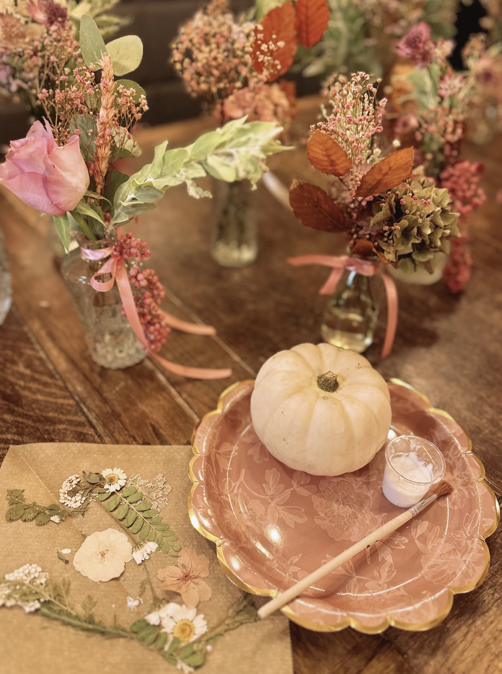 Decorative pumpkin on a pink floral plate surrounded by vases with pink and green floral arrangements on a wooden table, with dried flowers and a painted brush nearby.