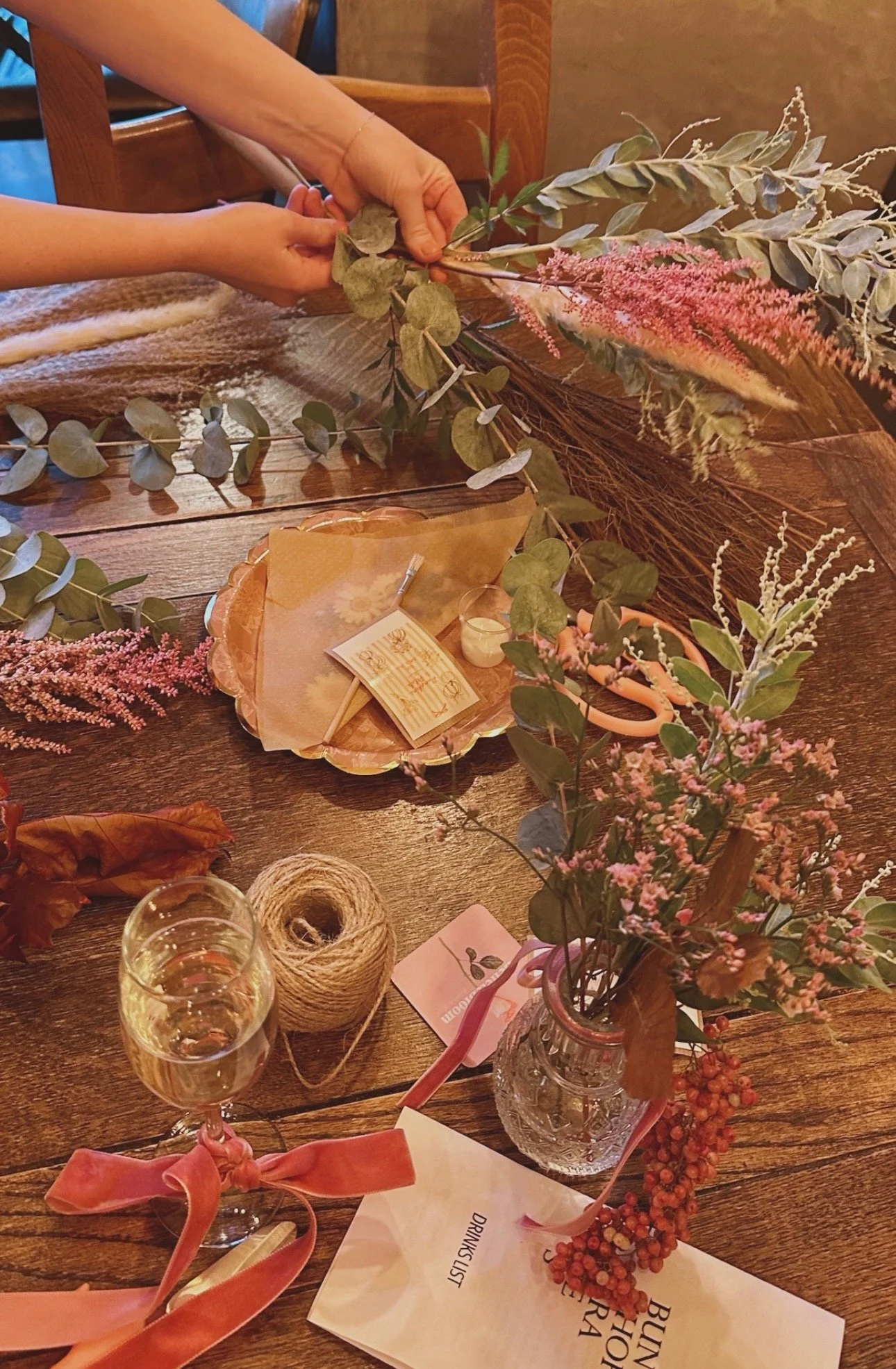 A wooden table decorated with pink and green floral arrangements, a glass of champagne with a pink ribbon, a spool of twine, a pair of pink scissors, a pink place card, and a pink plate with a note on it.