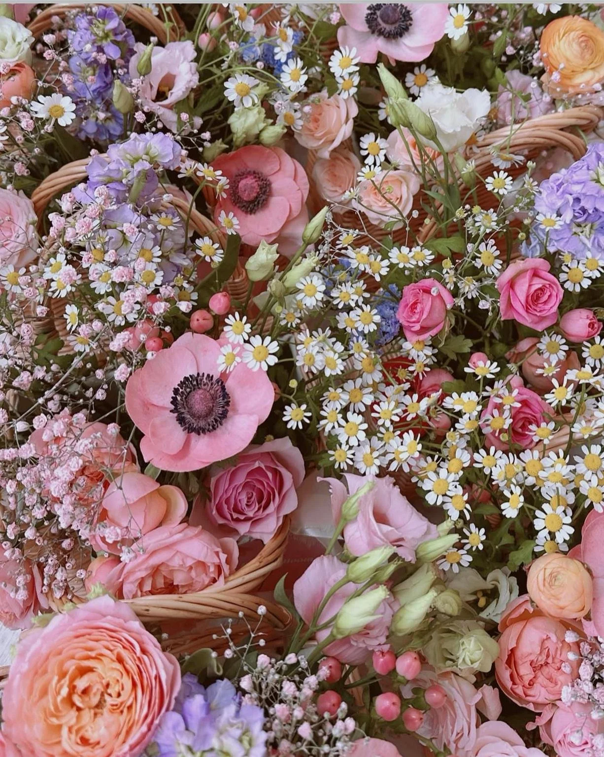 A close-up of a basket filled with a variety of pink, purple, white, and peach flowers including roses, anemones, chamomile daisies, and small blossoms.