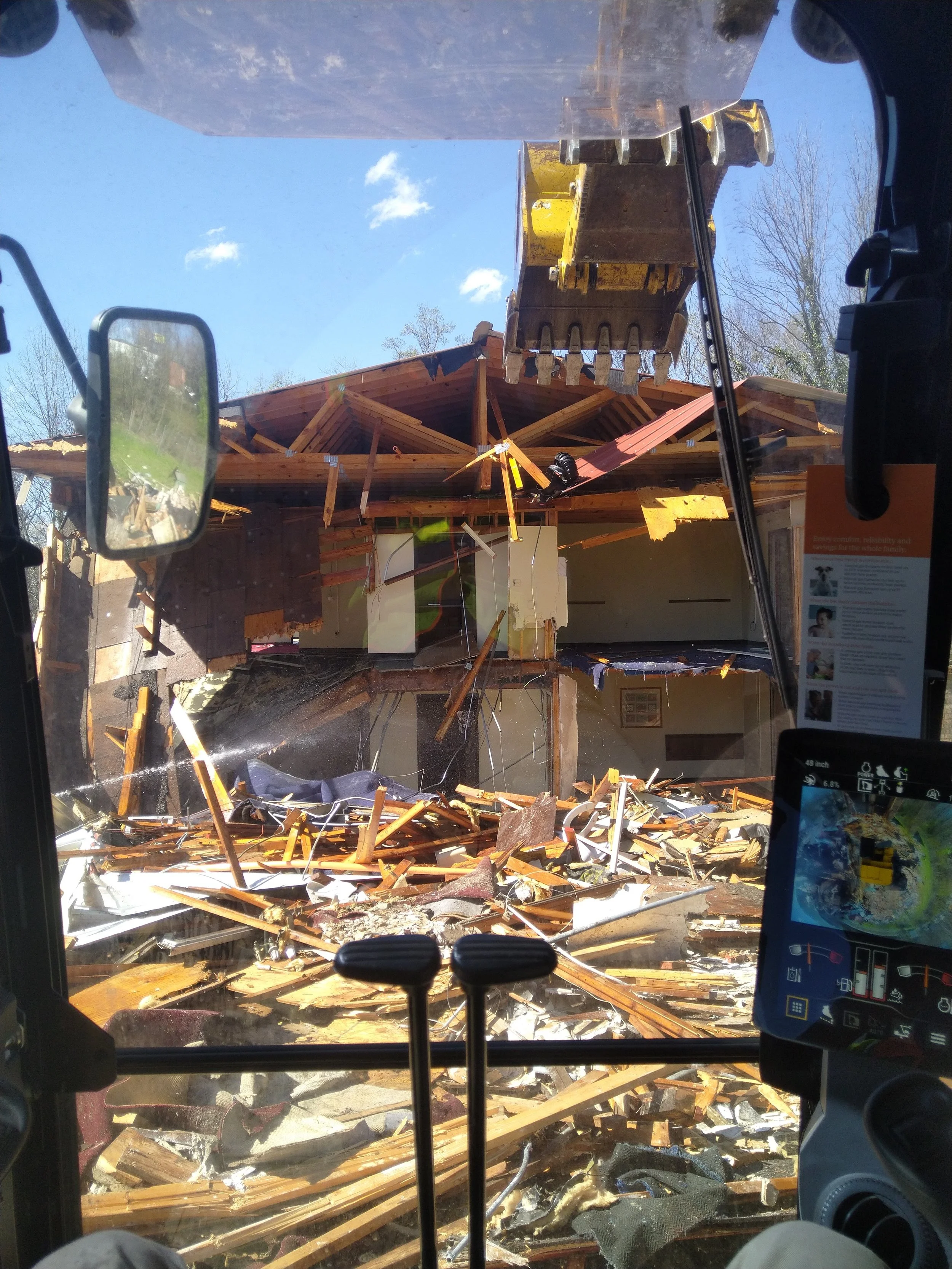 View from inside a construction vehicle looking at a house being demolished, with debris and broken wood scattered across the ground and part of the house's roof partially torn off.