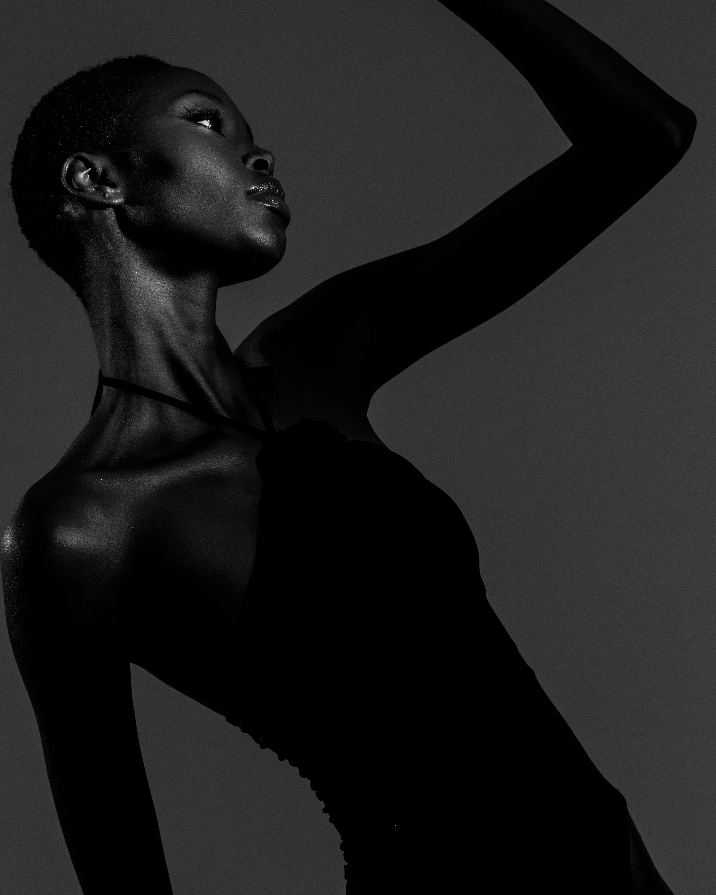 A black and white photo of a woman with short hair, wearing a sleeveless top and a choker necklace, raising her right arm with her hand above her head.