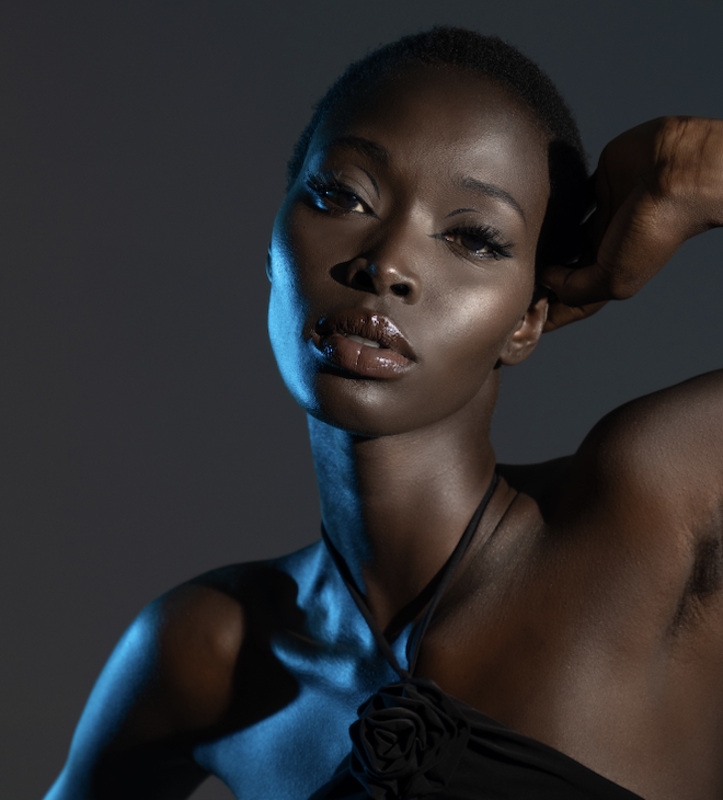A portrait of a confident woman with short hair, wearing a black strapless top, with one arm raised behind her head, and looking directly at the camera with a serious expression. The lighting creates a dramatic effect, highlighting her facial feature